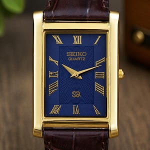 Op de afbeelding: Een rechthoekig Seiko Quartz polshorloge met een goudkleurige kast, een diepblauwe wijzerplaat met een golvend patroon en gouden Romeinse cijfers. Het horloge heeft goudkleurige wijzers en een donkerbruine leren band met krokodillenprint.
