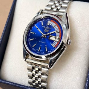 May include: A Seiko 5 automatic watch with a blue dial and a stainless steel strap. The watch has a date display and a striking orange and white detail on the dial. It is presented in a blue box with a white interior.