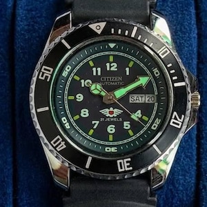 Può includere: Orologio Citizen Automatic con lunetta nera e cinturino in gomma nero. Il quadrante è verde scuro con dettagli bianchi e verdi luminosi. La data indica "SAB 20". L'orologio è in una scatola blu.