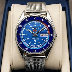 May include: A blue-dial Seiko automatic watch with a silver mesh strap. The watch face shows the day and date, with a 24-hour clock. The watch is presented in a blue box with a cream interior.