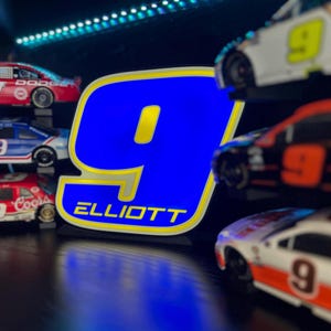 May include: A blue and yellow illuminated sign with the number 9 and the name "ELLIOTT" is the focal point. Miniature race cars with the number 9 are displayed on shelves, with red, white, and blue color schemes. The scene is set against a dark background.