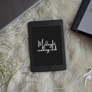 May include: A black Kindle e-reader displaying the text "Midnight reading club" on a white fluffy surface. A laptop, a book, and white earbuds are also present. The scene is lit with natural light.