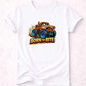 以下が含まれることがあります： 白いTシャツ。犬のような顔、茶色のボディ、青い車輪を持つ漫画のモンスタートラックのデザイン。テキスト「BORN TO BITE」は、トラックの下にカラフルで太字のフォントで表示されています。