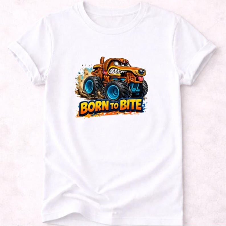 以下が含まれることがあります： 白いTシャツ。犬のような顔、茶色のボディ、青い車輪を持つ漫画のモンスタートラックのデザイン。テキスト「BORN TO BITE」は、トラックの下にカラフルで太字のフォントで表示されています。