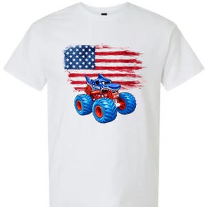 May include: White t-shirt featuring a shark-themed monster truck design with blue, red, and white colors. The design includes an American flag background, with the monster truck in the foreground.