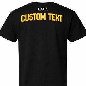 以下が含まれることがあります： 黒いTシャツ。上部に白い文字で「BACK」と書かれています。その下に、赤い縁取りのある黄色で「CUSTOM TEXT」という文字がプリントされています。このシャツは半袖で、クルーネックです。