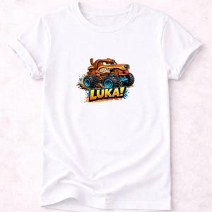 以下が含まれることがあります： 白いTシャツ。モンスターカーを運転する漫画の犬が描かれています。犬は茶色で怒った表情をしており、トラックは青い車輪をしています。「LUKA！」という名前がカラフルな文字でトラックの下に書かれています。