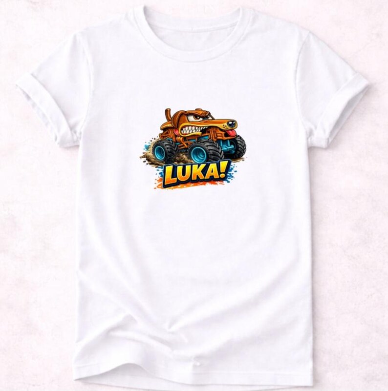以下が含まれることがあります： 白いTシャツ。モンスターカーを運転する漫画の犬が描かれています。犬は茶色で怒った表情をしており、トラックは青い車輪をしています。「LUKA！」という名前がカラフルな文字でトラックの下に書かれています。