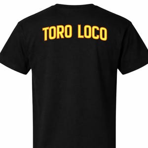 以下が含まれることがあります： 背中に黄色と赤の文字で「TORO LOCO」と書かれた黒いTシャツ。 このシャツはクルーネックです。