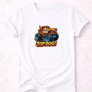 以下が含まれることがあります： 白いTシャツ。犬のカートゥーン・モンスタートラックのデザイン。トラックは茶色で、青い車輪と怒った表情をしています。「TOP DOG！」の文字がオレンジと青でトラックの下にあります。