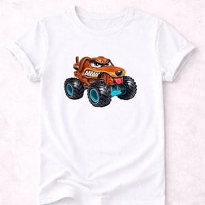 以下が含まれることがあります： 白いTシャツに、漫画のモンスタートラックのデザイン。トラックは茶色で、青色のアクセントと大きな黒いタイヤが付いています。犬は怒った表情をしており、舌を出しています。