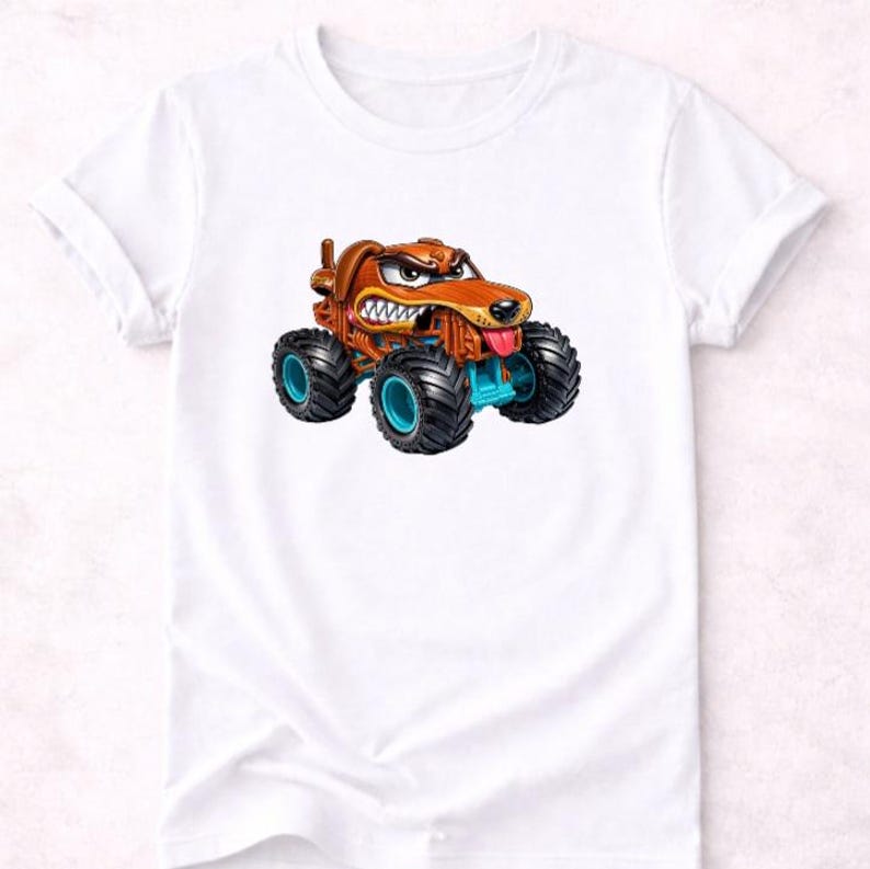 以下が含まれることがあります： 白いTシャツに、漫画のモンスタートラックのデザイン。トラックは茶色で、青色のアクセントと大きな黒いタイヤが付いています。犬は怒った表情をしており、舌を出しています。