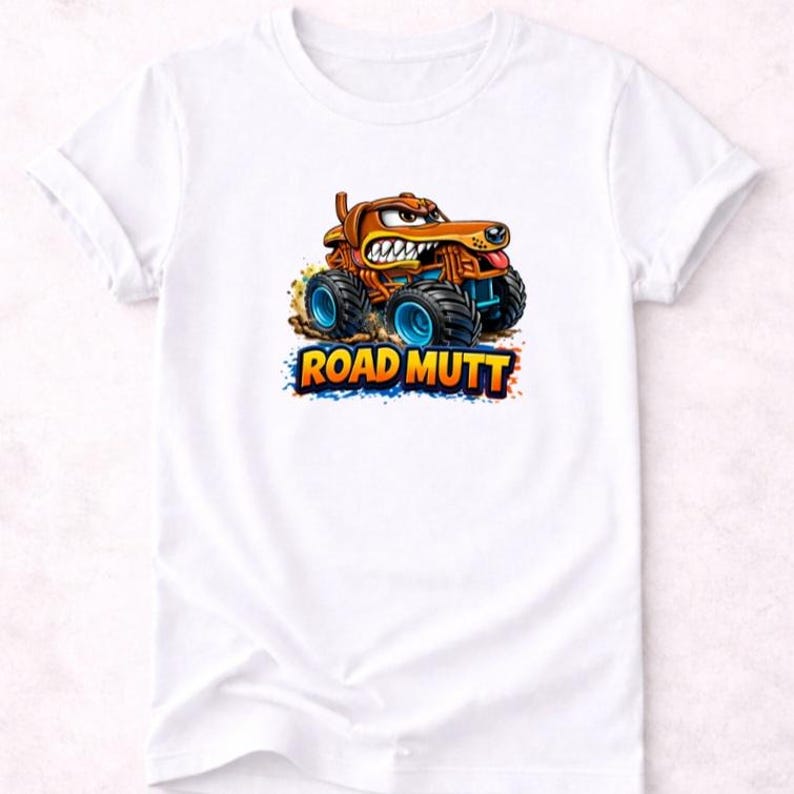 以下が含まれることがあります： 白いTシャツに、モンスターカーに乗った漫画の犬が描かれています。犬は茶色で、長い鼻と歯を見せた笑顔をしています。トラックは青い車輪で、「ROAD MUTT」の文字がカラフルなフォントで書かれています。