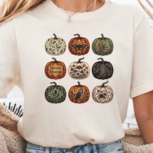 以下が含まれることがあります： クリーム色の半袖Tシャツで、9つの異なるカボチャがグリッド状に配置されたグラフィックプリントが施されています。各カボチャには、キノコ、フクロウ、蛾、葉、そして「Harvest Blessings」という文字など、秋をテーマにしたユニークなイラストが描かれています。