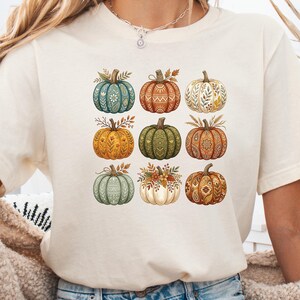 以下が含まれることがあります： クリーム色の半袖Tシャツで、3×3のグリッド状に配置された9つのカボチャのグラフィックプリントが施されています。それぞれのカボチャには、オレンジ、緑、ベージュの色合いで、花柄、幾何学模様、葉のモチーフなどのユニークなパターンが描かれています。