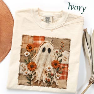 May include: An ivory t-shirt featuring a square patch design with a central ghost figure surrounded by embroidered orange and white flowers. The background of the patch consists of a patchwork pattern in shades of brown, tan, and orange plaid.