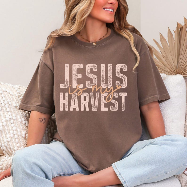 K&ouml;nnte beinhalten: Ein braunes T-Shirt mit kurzen &Auml;rmeln und dem Text "JESUS is my HARVEST" in einer hellen, abgenutzten Schriftart. Das Wort "is" ist in Schreibschrift mit einem kleinen Herzdetail gestaltet. Das Shirt hat einen Rundhalsausschnitt und eine lockere Passform.