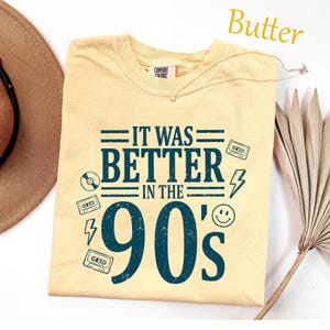May include: A butter yellow short-sleeved t-shirt featuring dark teal screen-printed text that reads IT WAS BETTER IN THE 90'S. The design includes graphic icons of cassette tapes, a lightning bolt, a smiley face, and a compact disc.