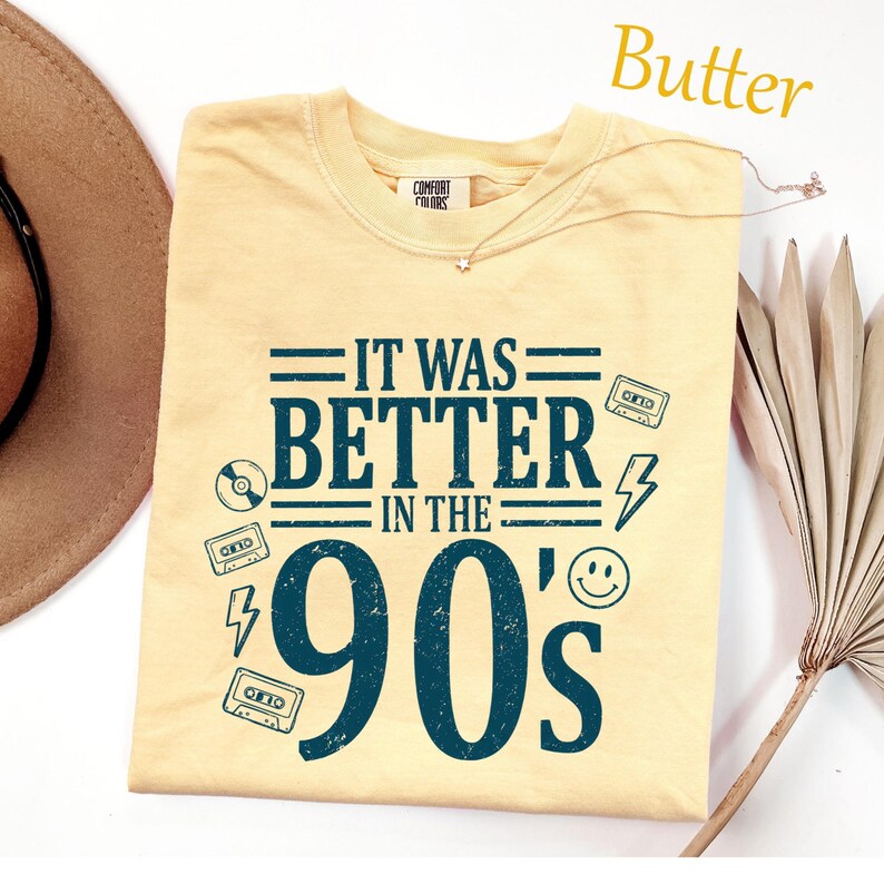 May include: A butter yellow short-sleeved t-shirt featuring dark teal screen-printed text that reads IT WAS BETTER IN THE 90'S. The design includes graphic icons of cassette tapes, a lightning bolt, a smiley face, and a compact disc.