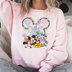 May include: A light pink sweatshirt featuring a colorful graphic of Disney characters, including Mickey and Minnie Mouse, Donald Duck, and Goofy, in front of a castle with fireworks. The design is framed by a Mickey Mouse ear outline.