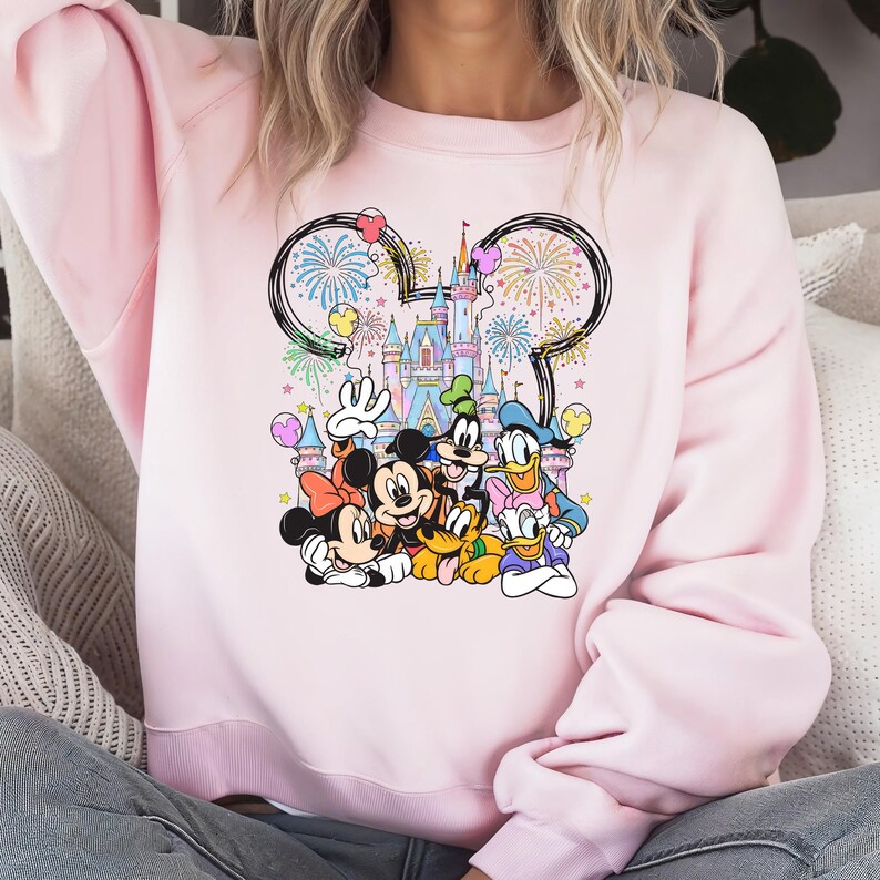 May include: A light pink sweatshirt featuring a colorful graphic of Disney characters, including Mickey and Minnie Mouse, Donald Duck, and Goofy, in front of a castle with fireworks. The design is framed by a Mickey Mouse ear outline.
