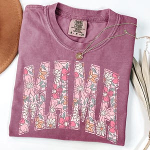 May include: A mauve short-sleeved t-shirt featuring the word MAMA in large block letters filled with a floral pattern of pink, orange, and white flowers with green leaves. The shirt has a crew neckline and a Comfort Colors brand label inside.
