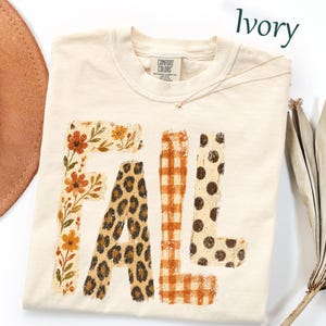 May include: An ivory t-shirt with the word FALL printed in large letters. The F features orange flowers and green leaves, the A has a brown leopard print, the L displays an orange gingham pattern, and the second L has brown polka dots.
