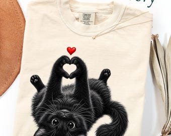 May include: An ivory-colored crew neck t-shirt featuring a graphic of a black cat lying on its back. The cat has its front paws raised to form a heart shape, with a small red heart icon positioned directly above its paws. The shirt has a Comfort Colors label.