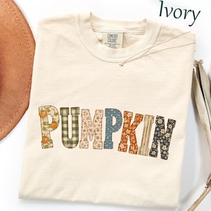 May include: An ivory crew neck t-shirt with the word PUMPKIN printed across the chest. Each letter features a different pattern, including pumpkins, green plaid, autumn leaves, blue dots, orange floral, stripes, and dark floral designs.