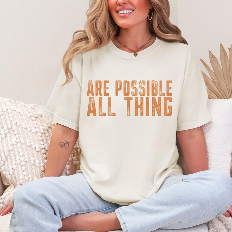 K&ouml;nnte beinhalten: Ein cremefarbenes Kurzarm-T-Shirt mit dem orangefarbenen Text ARE POSSIBLE ALL THING, der in einer abgenutzten Blockschrift auf der Brust gedruckt ist. Das T-Shirt hat einen Rundhalsausschnitt und eine lockere Passform, kombiniert mit einer hellblauen Jeans.