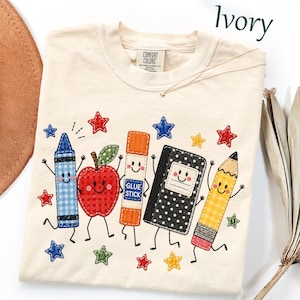 May include: An ivory t-shirt featuring embroidered school supply characters including a blue gingham crayon, a red gingham apple, an orange glue stick, a black polka-dot notebook, and a yellow pencil, all surrounded by colorful stars.