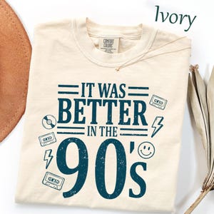 May include: An ivory t-shirt featuring dark blue text that reads IT WAS BETTER IN THE 90's. The design includes graphic illustrations of cassette tapes, a lightning bolt, a smiley face, and a compact disc, all arranged around the central text.