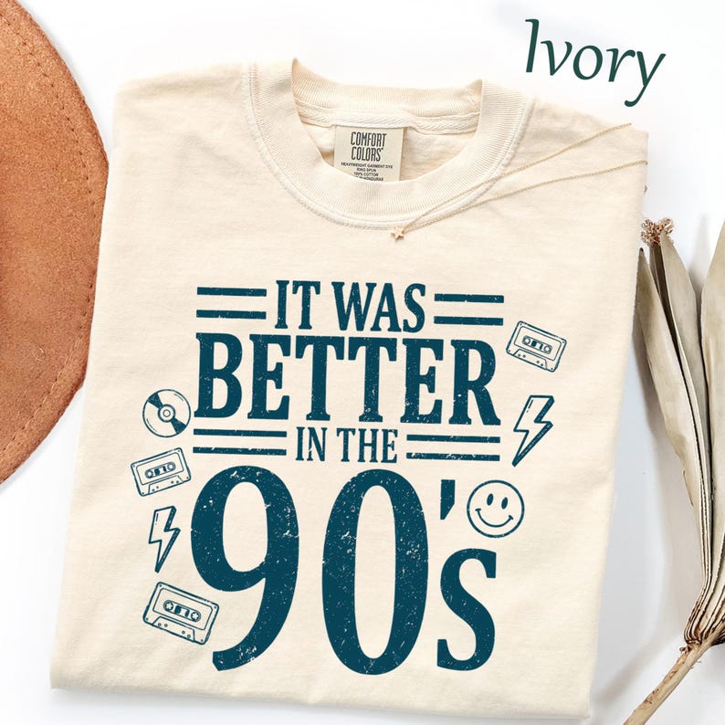 May include: An ivory t-shirt featuring dark blue text that reads IT WAS BETTER IN THE 90's. The design includes graphic illustrations of cassette tapes, a lightning bolt, a smiley face, and a compact disc, all arranged around the central text.
