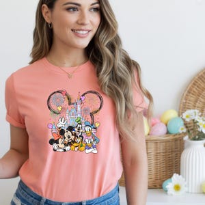 May include: Coral-colored t-shirt featuring a graphic of Mickey Mouse, Minnie Mouse, Donald Duck, Daisy Duck, and Goofy in front of a castle. The design includes fireworks and balloons. The shirt has short sleeves.