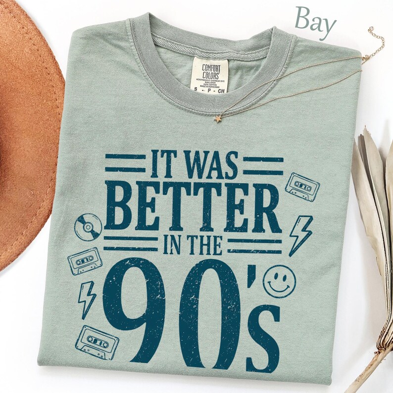 May include: A sage green crew neck t-shirt featuring dark blue distressed text that reads IT WAS BETTER IN THE 90's. The design includes graphic icons of cassette tapes, lightning bolts, a smiley face, and a compact disc. The shirt has a Comfort Colors label.