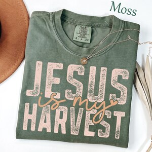 K&ouml;nnte beinhalten: Ein moosgr&uuml;nes Kurzarm-T-Shirt mit Rundhalsausschnitt und einem Comfort Colors-Etikett. Das Shirt zeigt den Schriftzug "JESUS IS MY HARVEST" in einer verwaschenen hellrosa und beigen Schrift. Eine d&uuml;nne goldfarbene Halskette mit kleinem Anh&auml;nger liegt auf dem Shirt.