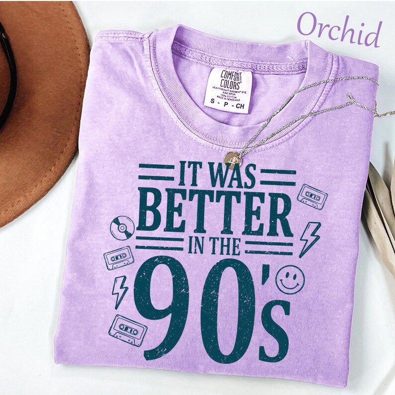 May include: An orchid-colored short-sleeve t-shirt with a dark teal graphic print that reads IT WAS BETTER IN THE 90'S. The design includes illustrations of cassette tapes, a lightning bolt, a smiley face, and a compact disc. A silver chain necklace rests on the collar.