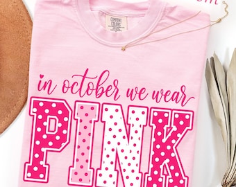 May include: A light pink t-shirt featuring the text "in october we wear" in a cursive script above the word "PINK" in large block letters. The letters are filled with a white and pink polka dot pattern. The shirt has a crew neckline.