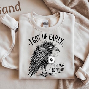 May include: A sand-colored crewneck sweatshirt featuring a black graphic of a grumpy crow holding a steaming coffee mug. The text reads, I GOT UP EARLY, THERE WAS NO WORM. The garment has a Gildan brand label at the inner neckline.