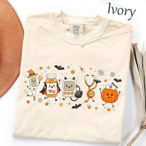 May include: An ivory t-shirt featuring a graphic print of cartoon medical items dressed for Halloween, including a thermometer in a witch hat, a pill bottle in a cape, a blood glucose monitor, a stethoscope, and a pumpkin wearing a nurse cap.