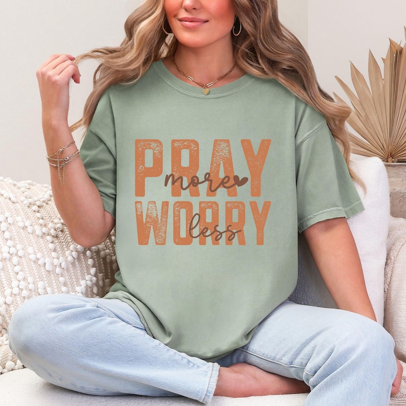 K&ouml;nnte beinhalten: Ein salbeigr&uuml;nes Kurzarm-T-Shirt mit dem orangefarbenen und braunen Text PRAY more WORRY less. Das T-Shirt hat einen Rundhalsausschnitt und eine lockere Passform, wobei die W&ouml;rter PRAY und WORRY in einer fetten, abgenutzten Schriftart sind.