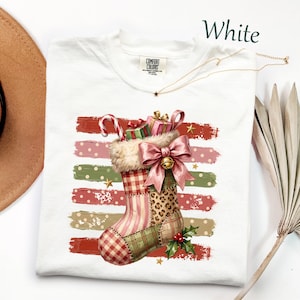 May include: A white t-shirt with a Comfort Colors label featuring a graphic of a Christmas stocking with a pink bow, candy cane, and gifts. The stocking has plaid, striped, and leopard print patterns, set against horizontal brush strokes.