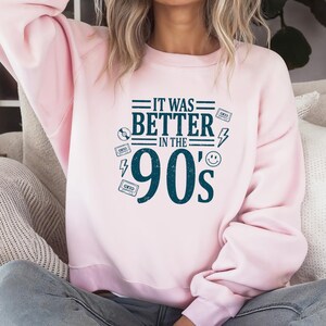 May include: A light pink crewneck sweatshirt featuring dark blue text that reads IT WAS BETTER IN THE 90'S. The design includes graphic elements such as a cassette tape, a compact disc, a lightning bolt, and a smiley face icon surrounding the text.