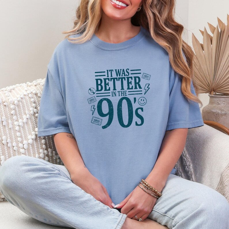 May include: A light blue crewneck t-shirt featuring dark teal text that reads IT WAS BETTER IN THE 90'S. The graphic includes small illustrations of cassette tapes, a lightning bolt, a smiley face, and a compact disc surrounding the large text.
