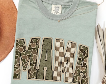 May include: A sage green short-sleeve t-shirt featuring the word MAMA in large block letters. The letters M, M, and A are filled with a camouflage pattern, while the letter A features a brown and white checkered pattern.