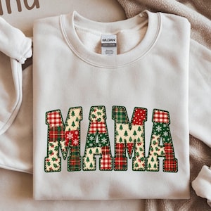 May include: A light beige crewneck sweatshirt featuring the word MAMA in large block letters. Each letter is filled with a patchwork pattern of red, green, and white holiday motifs including plaid, Christmas trees, snowflakes, and holly berries.