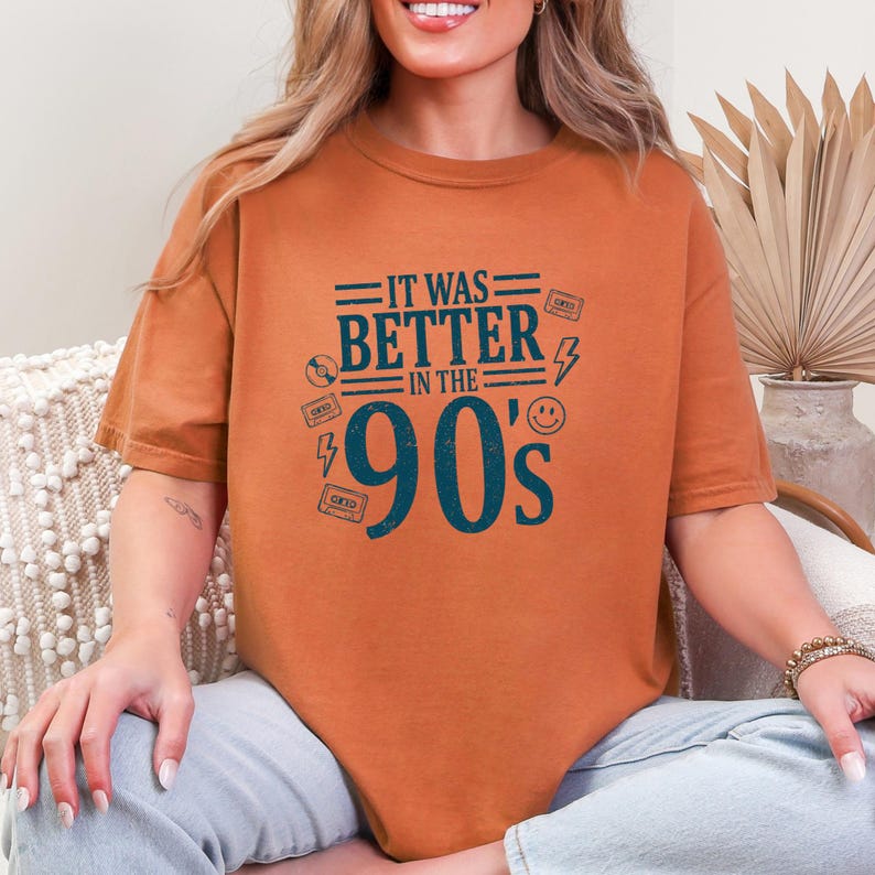 May include: A burnt orange short-sleeve t-shirt featuring dark teal text that reads IT WAS BETTER IN THE 90's. The graphic includes small illustrations of a compact disc, cassette tapes, a lightning bolt, and a smiley face surrounding the text.