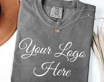 May include: A charcoal gray short-sleeve t-shirt with a crew neckline and a Comfort Colors brand label at the inner collar. The shirt features the white cursive text "Your Logo Here" printed across the chest and includes a thin silver necklace.