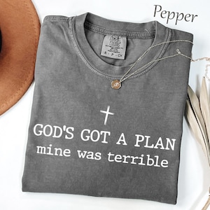 May include: A charcoal gray short-sleeve t-shirt with a crew neckline. The shirt features a white graphic of a cross above the text "GOD'S GOT A PLAN mine was terrible". A thin silver-toned chain necklace with a small circular pendant rests on the shirt.