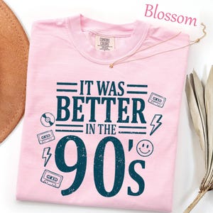 May include: A light pink t-shirt featuring dark blue distressed text that reads IT WAS BETTER IN THE 90'S. The graphic includes illustrations of cassette tapes, a compact disc, lightning bolts, and a smiley face. The shirt has a crew neckline.